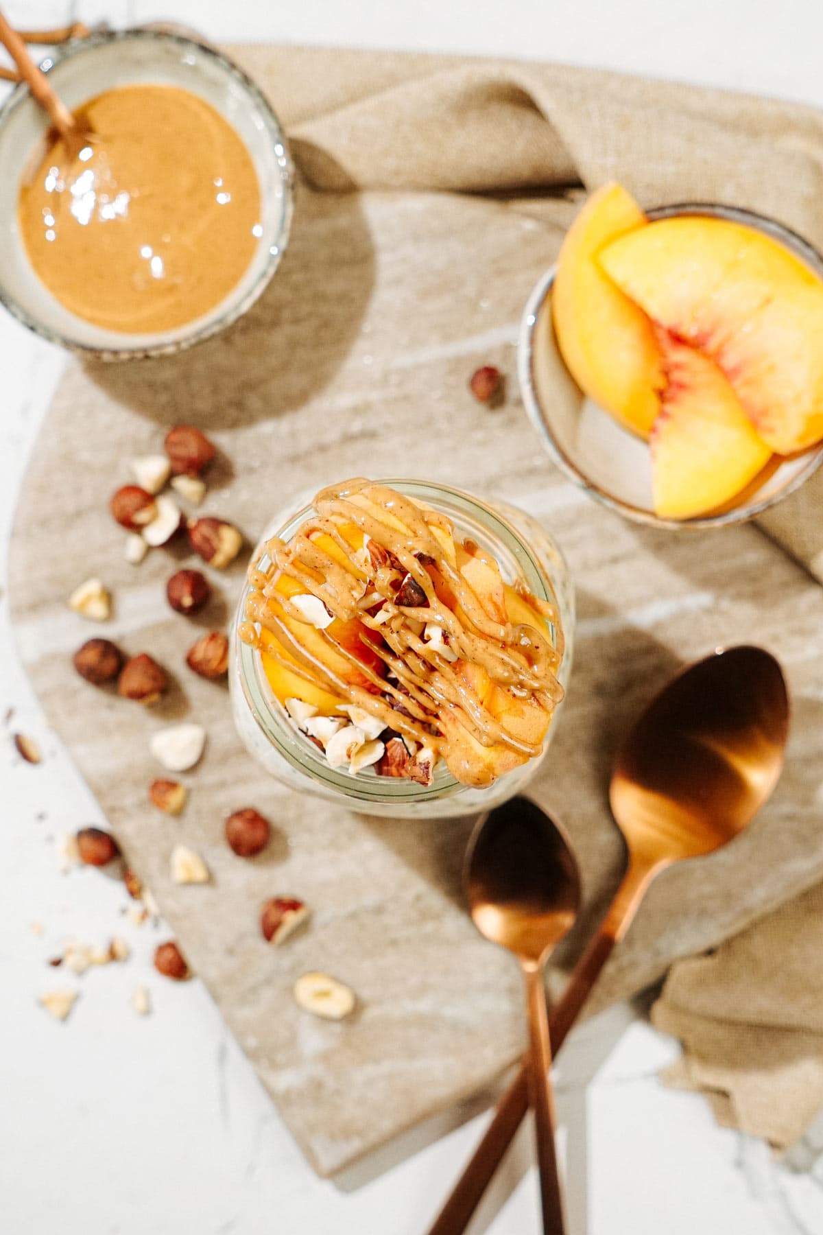 Bird's-eye view of the Mason jar with overnight oats is on the marble cut board. There are also some hazelnuts, two bronze spoons, a tiny bowl with peanut butter and a small dish with some slices of peach | Hurry The Food Up