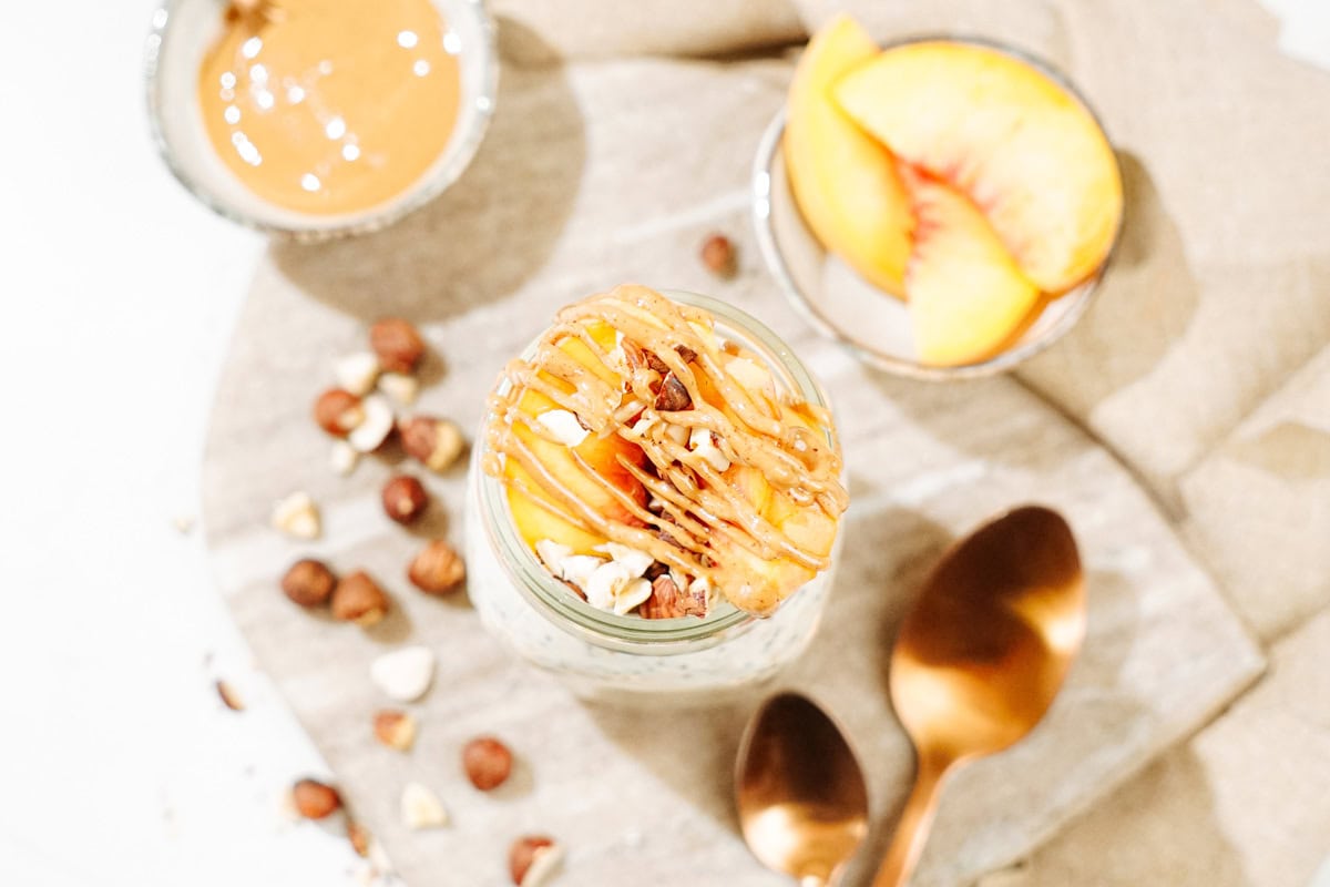Bird's-eye view of the Mason jar with overnight oats is on the marble cut board. There are also some hazelnuts, two bronze spoons, a tiny bowl with peanut butter and a small dish with some slices of peach | Hurry The Food Up