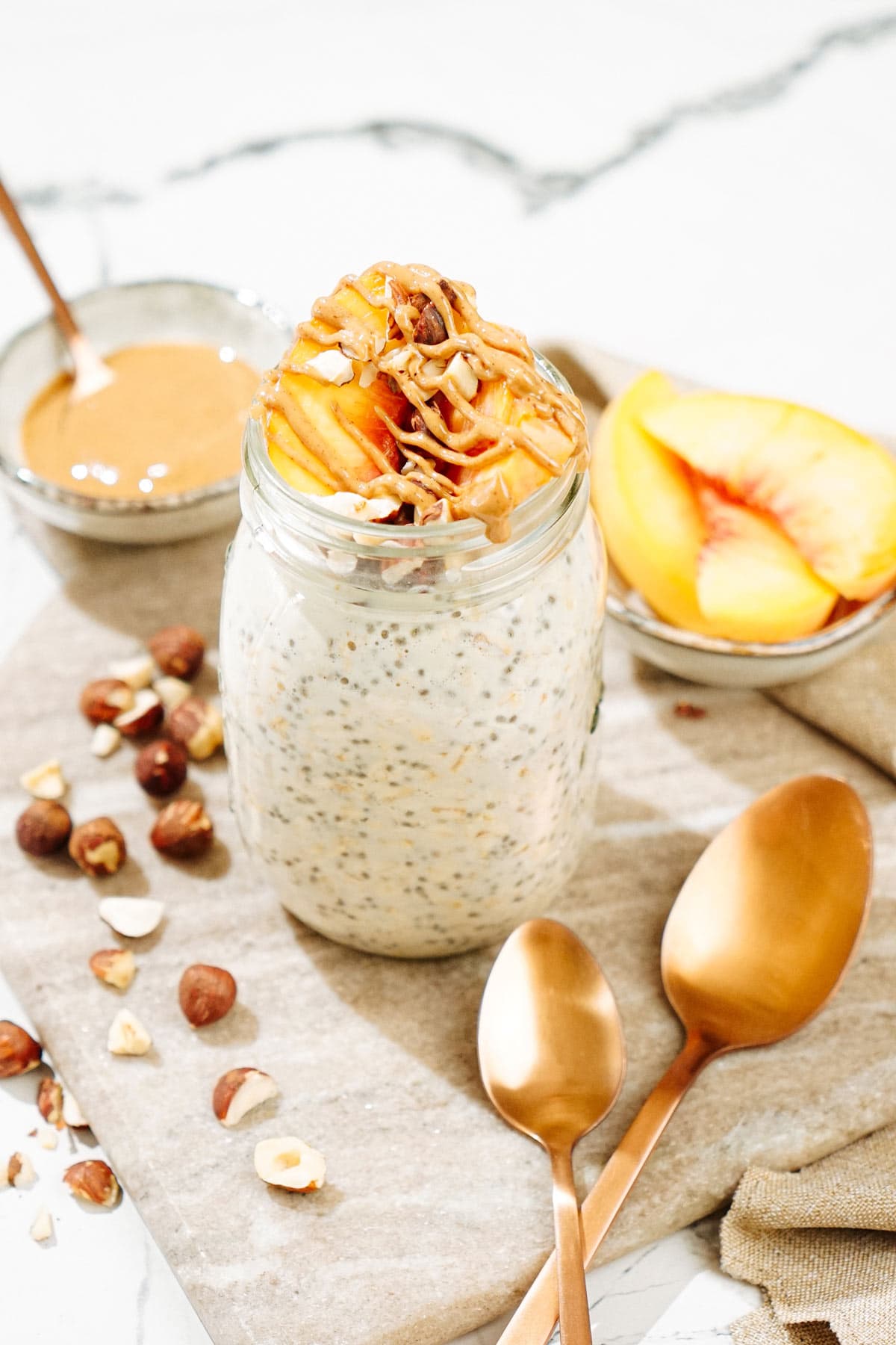The Mason jar with overnight oats is on the marble surface. There are some hazelnuts, two bronze spoons, a tiny bowl with peanut butter and a small dish with some slices of peach | Hurry The Food Up