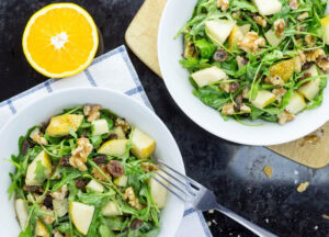 Two white bowls of pear and walnut salad are placed on a cutting board and cloth over a table with an orange and crumbs of walnuts | Hurry The Food Up