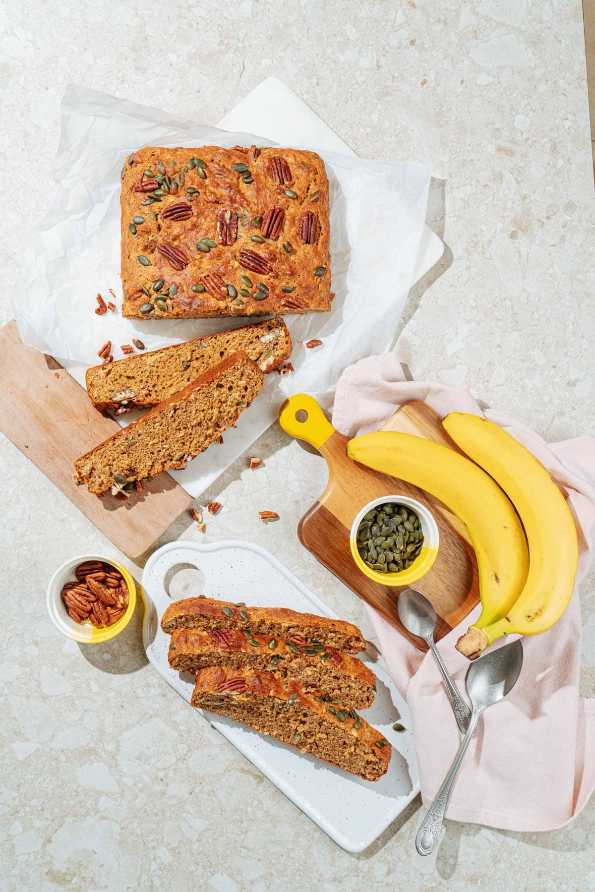 Bird's-eye view of protein banana bread that is on the white board. Around it, there are small bowls with pumpkin seeds and pecans, a spoon, bananas, and other pieces of banana bread | Hurry The Food Up