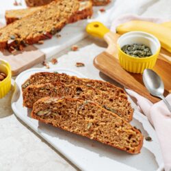The three pieces of deeply golden brown protein banana bread lie on the white board. Around it, there are small bowls with pumpkin seeds and pecans, a spoon, a banana, and other pieces of banana bread | Hurry The Food Up