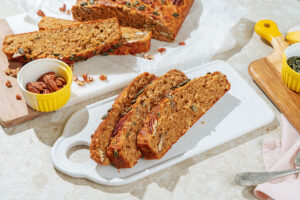 The three pieces of deeply golden brown protein banana bread lie on the white board. Around it, there are small bowls with pumpkin seeds and pecans, a spoon, and other pieces of banana bread | Hurry The Food Up