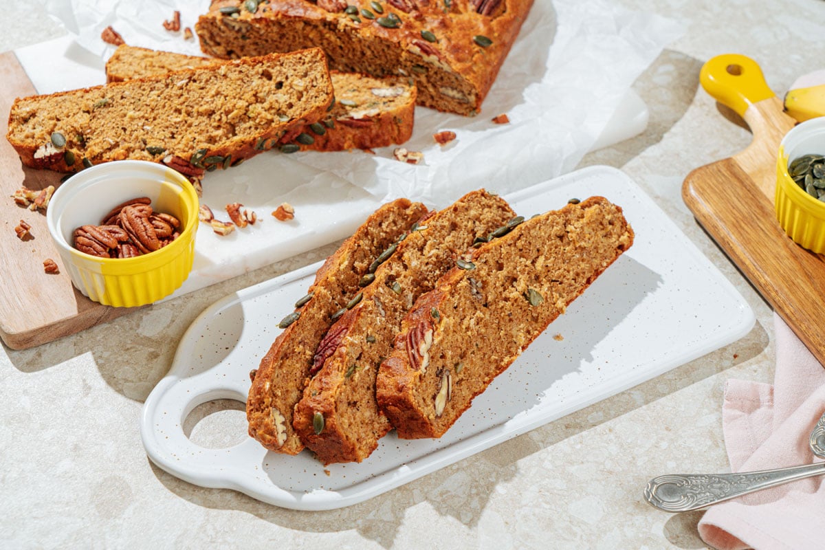 The three pieces of deeply golden brown protein banana bread lie on the white board. Around it, there are small bowls with pumpkin seeds and pecans, a spoon, and other pieces of banana bread | Hurry The Food Up
