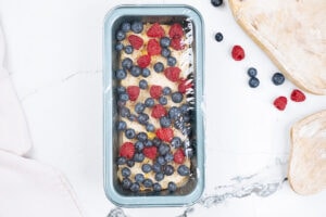 A loaf tin with protein French toast covered by food wrap is on the light surface. Next to it, there are some berries and wooden dishes | Hurry The Food Up
