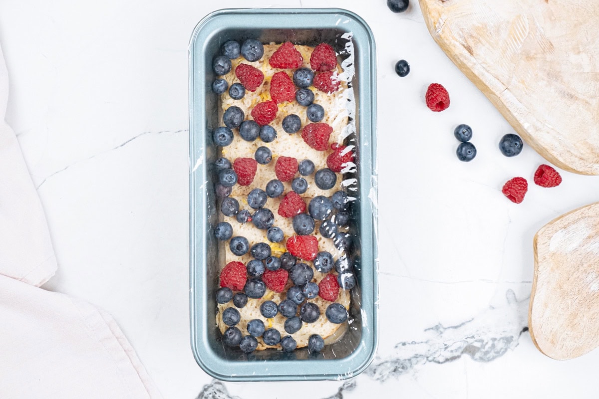 A loaf tin with protein French toast covered by food wrap is on the light surface. Next to it, there are some berries and wooden dishes | Hurry The Food Up