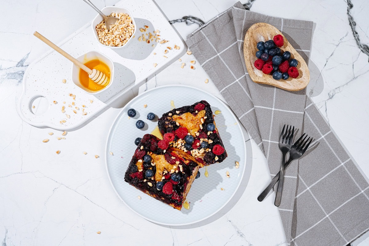 Bird's-eye view of a nice white plate with French toast is on a light surface. Next to it, there are grey napkin, two forks, a white tray with honey and chopped nuts, and a wooden plate with berries | Hurry The Food Up