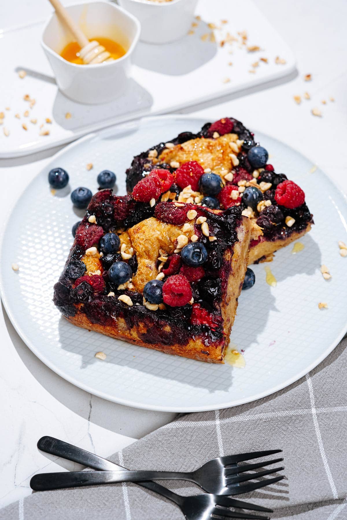 A white plate with French toast is on a light surface. Next to it, there are grey napkin, two forks and a white tray with honey and chopped nuts | Hurry The Food Up