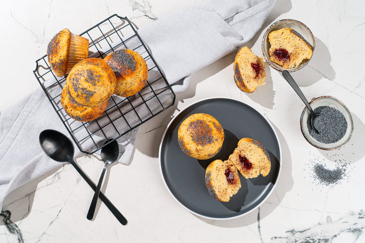 Bird's-eye view of protein muffins are on the dark plate. Near it there are some whole muffins on a metal grid and a small bowl with poppy seeds etc | Hurry The Food Up