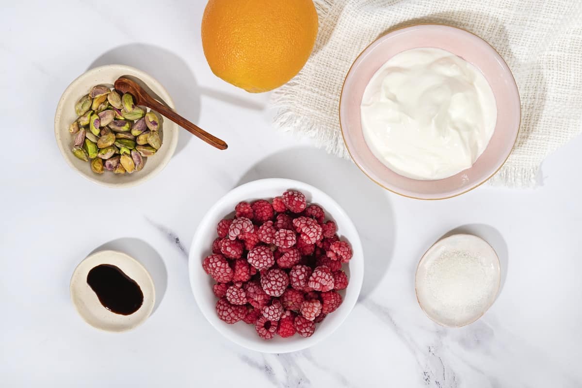 An orange, a bowl of frozen raspberries, pistachios, yogurt, sugar and syrup on a marble surface | Hurry The Food Up