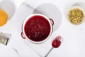 A pan full of raspberry compote, a spoon of compote next to it, a small grater and an orange to the left. | Hurry The Food Up