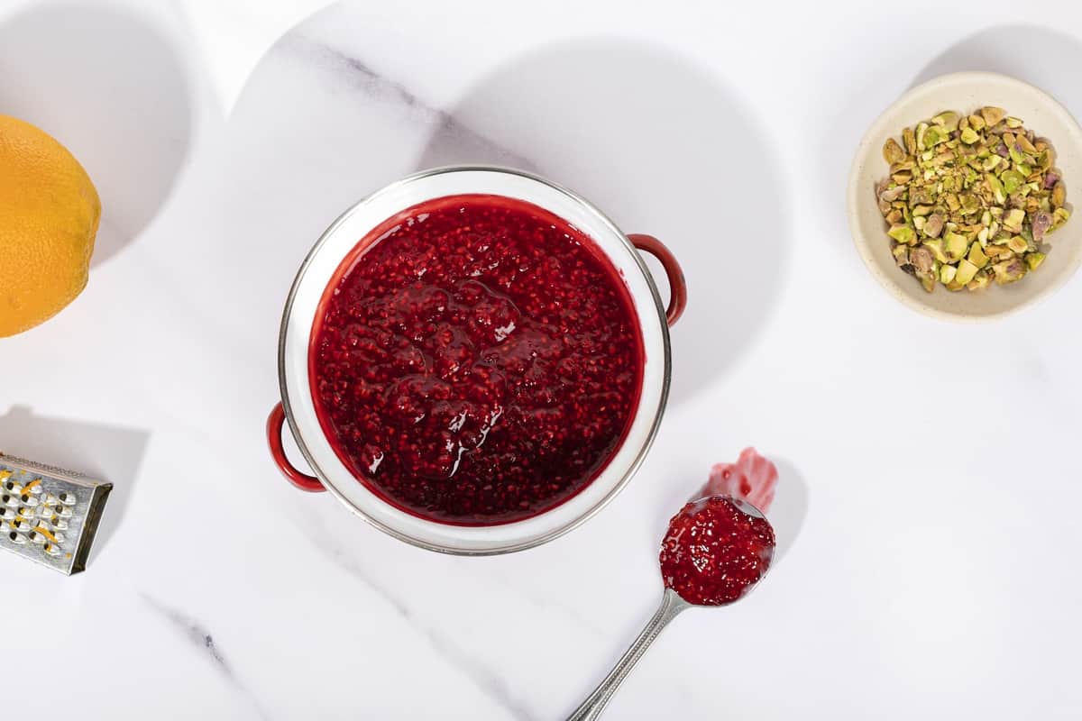 A pan full of raspberry compote, a spoon of compote next to it, a small grater and an orange to the left. | Hurry The Food Up