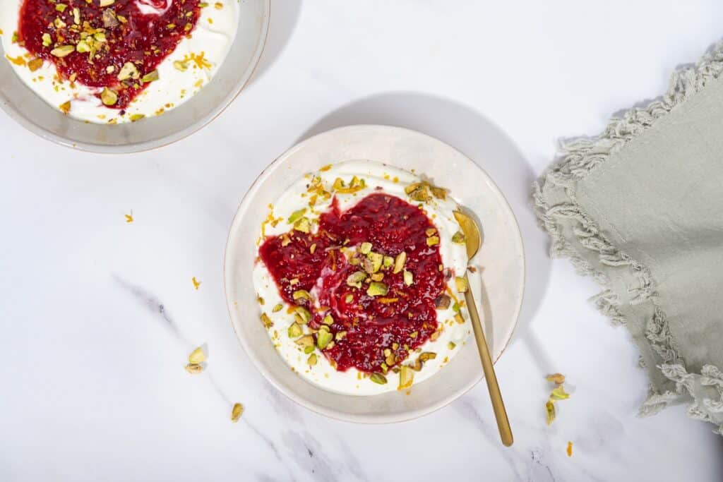 2 white bowls of yogurt, compote and pistachios, one with a golden spoon in it, on a marble surface. | Hurry The Food Up