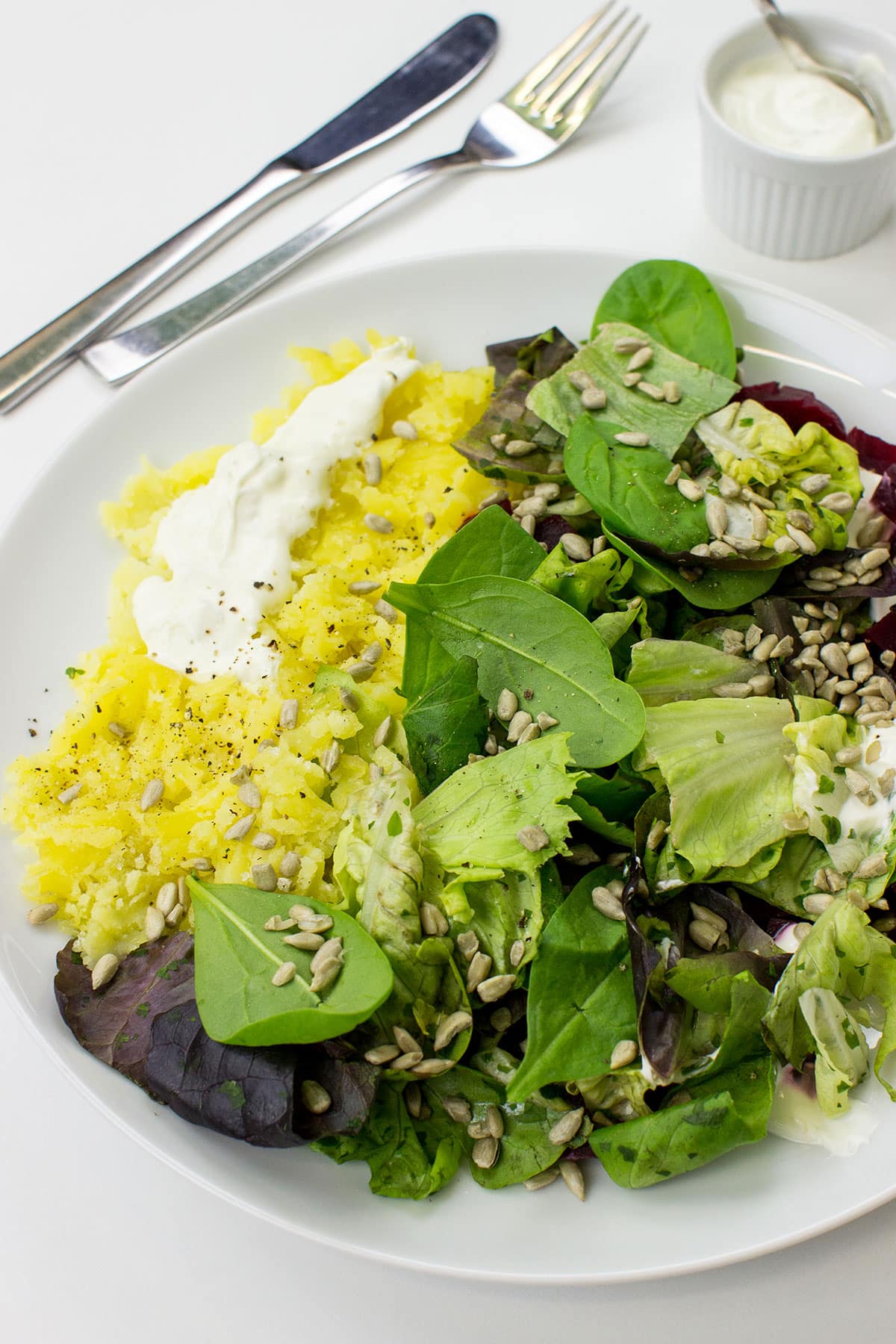 A white plate with Roasted Beetroot & Potato Salad, featuring yellow potatoes, red beetroot cubes, mixed greens, sunflower seeds, and dollops of lime crème fraîche, accompanied by a small bowl of crème fraîche, a knife, and a fork on a white surface.