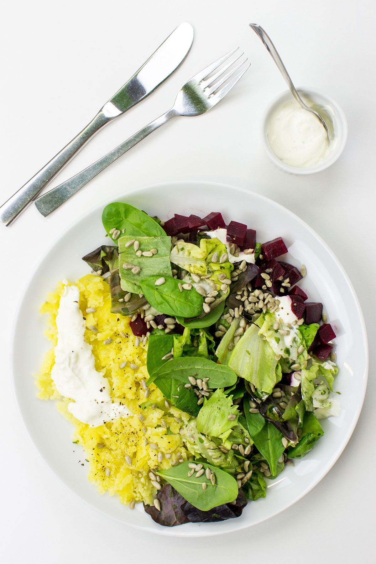 A white plate with Roasted Beetroot & Potato Salad, featuring yellow potatoes, red beetroot cubes, mixed greens, sunflower seeds, and dollops of lime crème fraîche, accompanied by a small bowl of crème fraîche, a knife, and a fork on a white surface.