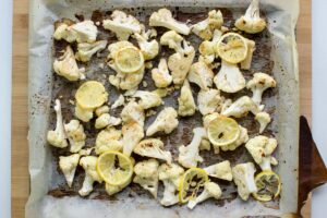 There is a baking sheet covered with parchment paper with baked cauliflower and lemon on a wooden surface | Hurry The Food Up