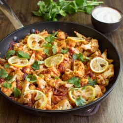 Roasted Cauliflower Curry is in the pan on the wooden surface. Next to it there is a plate with yogurt and fresh sprig cilantro | Hurry The Food Up