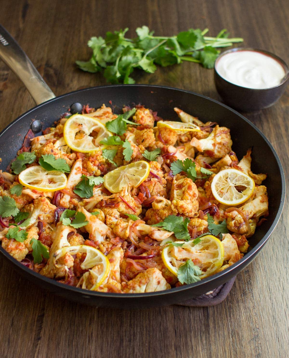 Roasted Cauliflower Curry is in the pan on the wooden surface. There is a bowl with yogurt and fresh sprig cilantro next to it | Hurry The Food Up