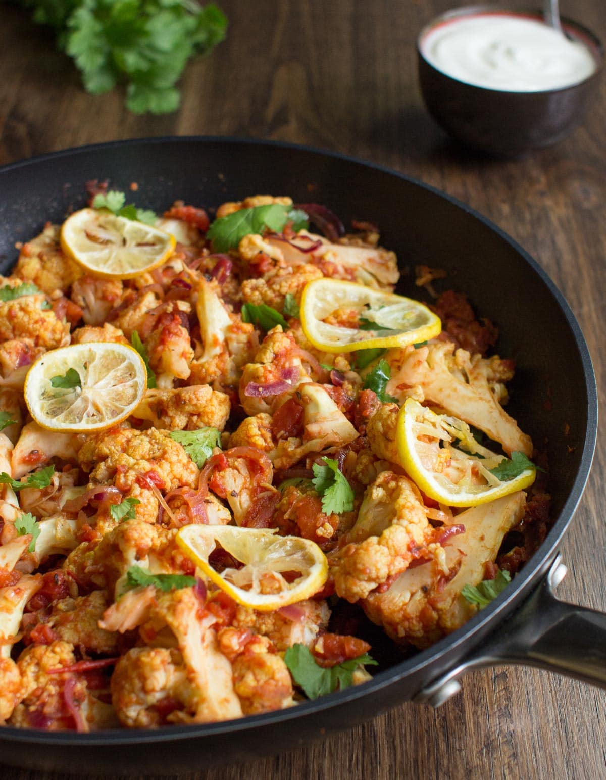 Roasted Cauliflower Curry is in the pan on the wooden surface. Next to it there is a bowl with yogurt and fresh sprig cilantro | Hurry The Food Up
