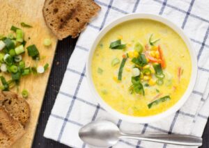 A close up view of a bowl of vegetarian corn chowder on a checked tea towel. There is a spoon beside it | Hurry The Food Up
