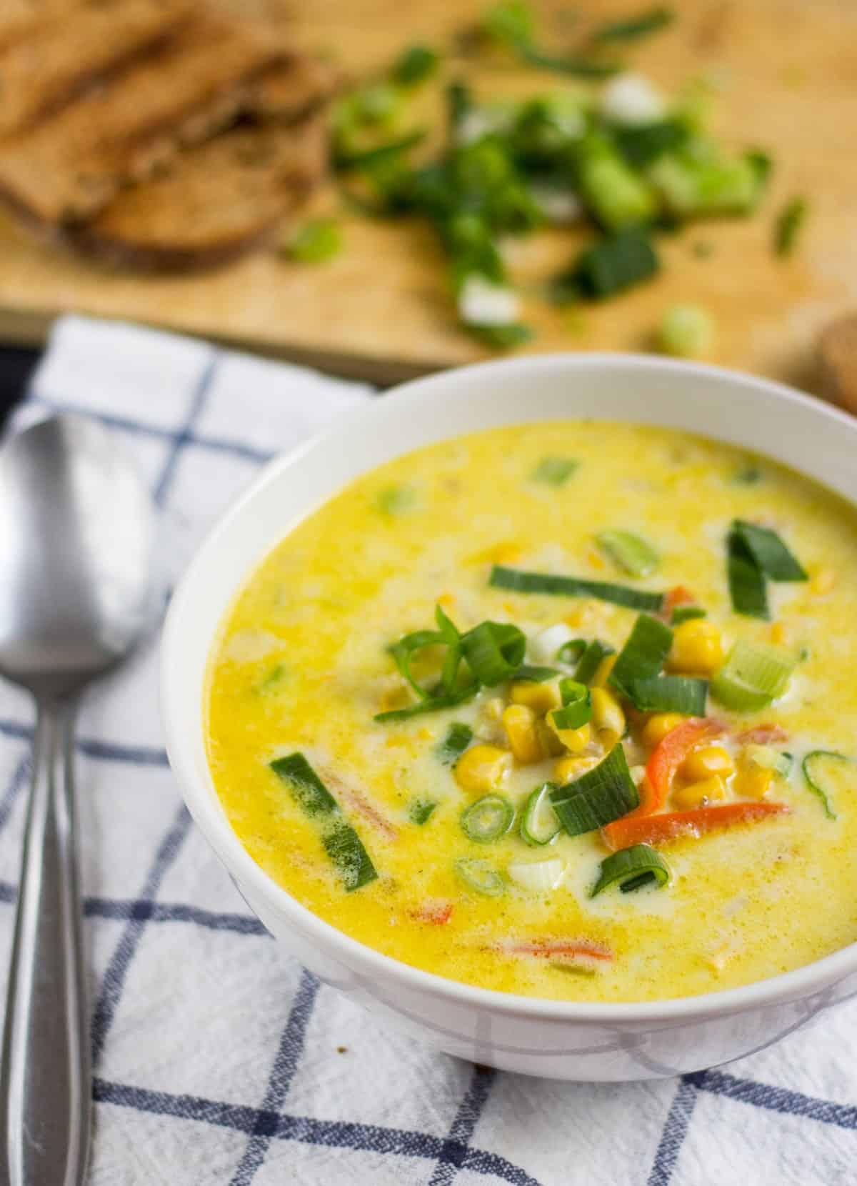A close up view of a bowl of vegetarian corn chowder on a checked tea towel. There is a spoon beside it | Hurry The Food Up