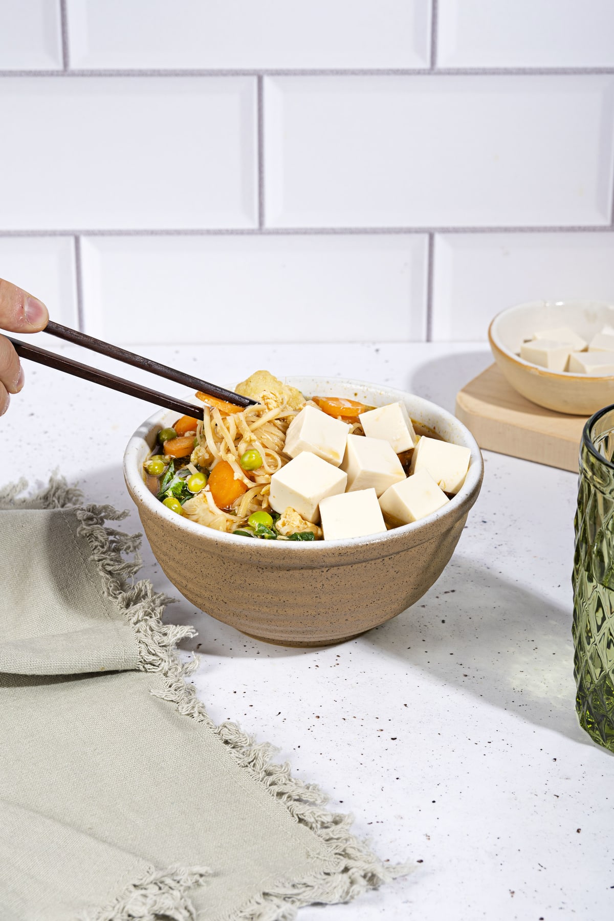 A person is picking up noodles from the bowl of egg drop soup that is on the white table along the bowl with tofu cubes,  grey towel and a glass of water | Hurry The Food Up