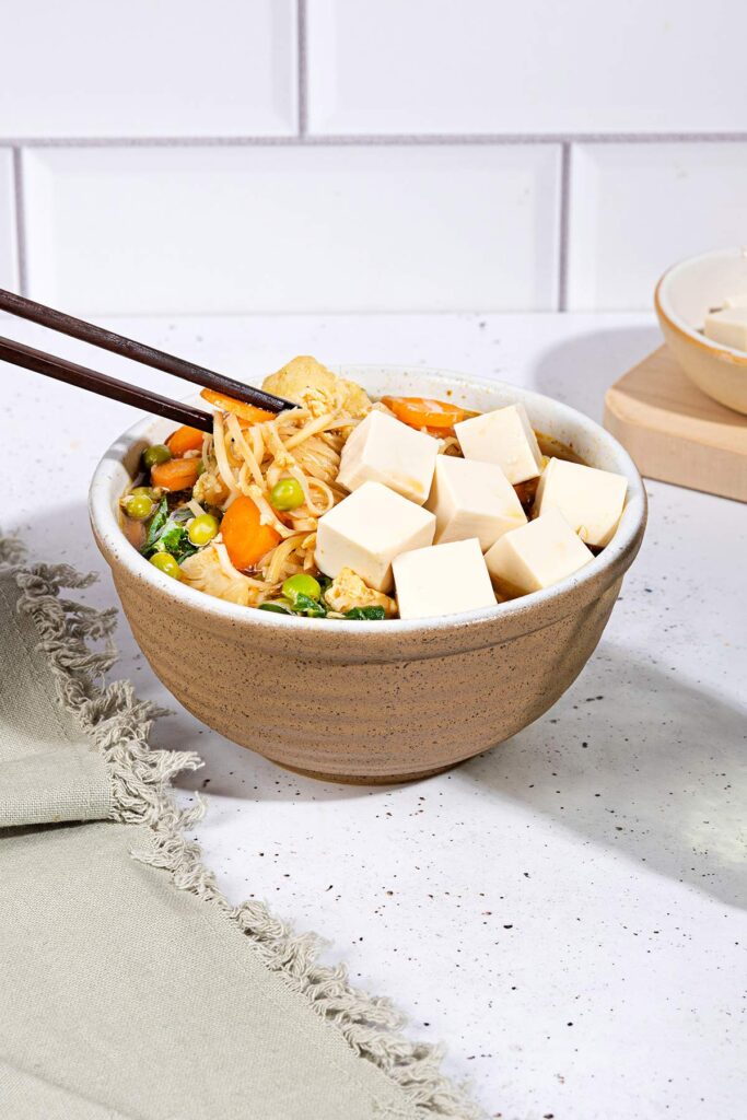 A person is picking up noodles from the bowl of egg drop soup that is on the white table along the bowl with tofu cubes, grey towel and a glass of water | Hurry The Food Up