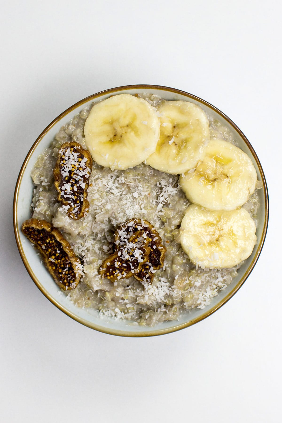 A bowl of tropical quinoa breakfast porridge topped with banana slices, dried fig pieces, and shredded coconut on a white background.