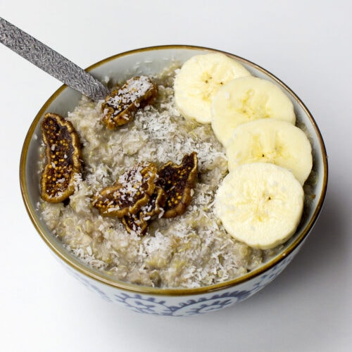 A bowl of tropical quinoa breakfast porridge topped with banana slices, dried fig pieces, and shredded coconut, served with a silver spoon on a white background.