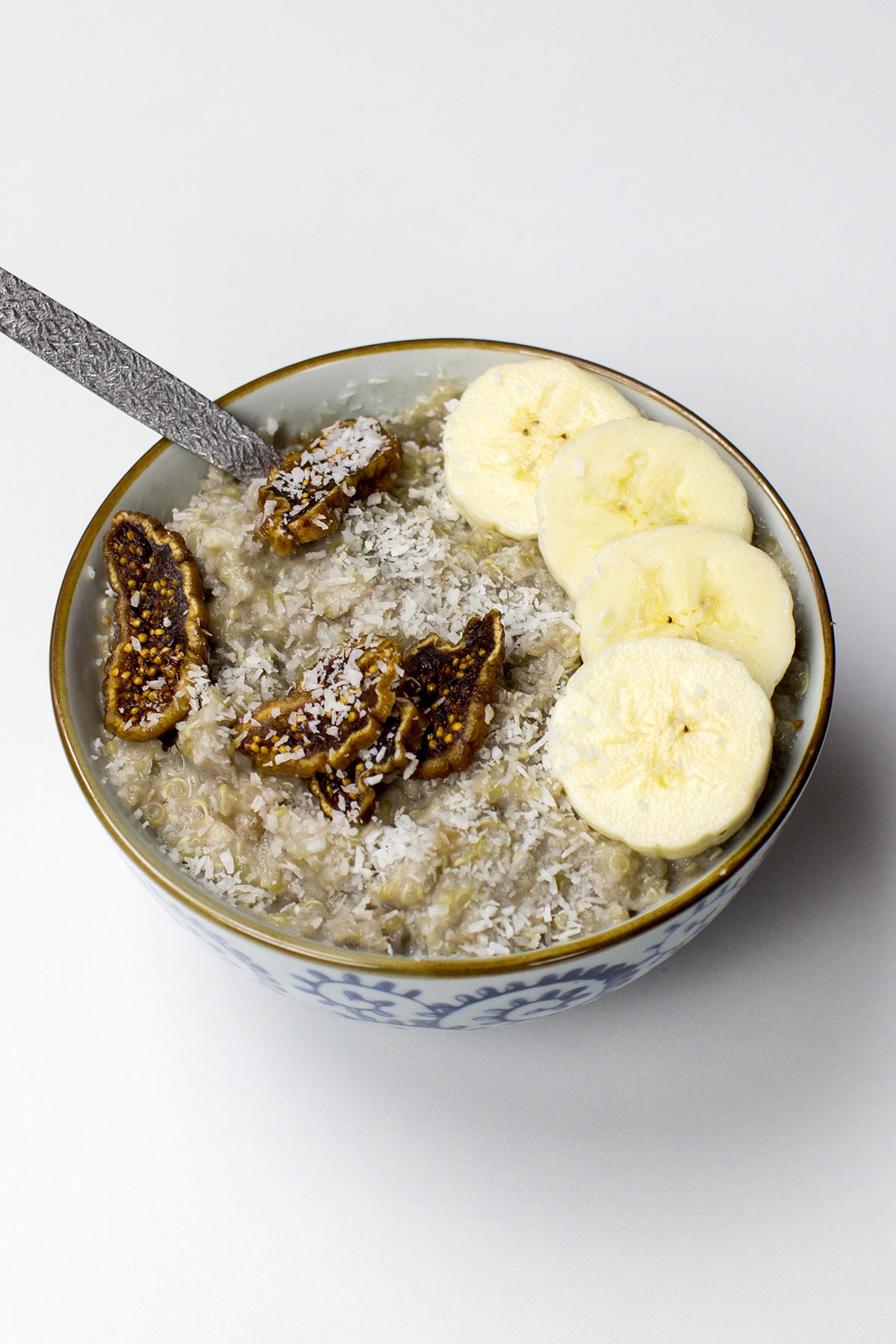 A bowl of tropical quinoa breakfast porridge topped with banana slices, dried fig pieces, and shredded coconut, served with a silver spoon on a white background.