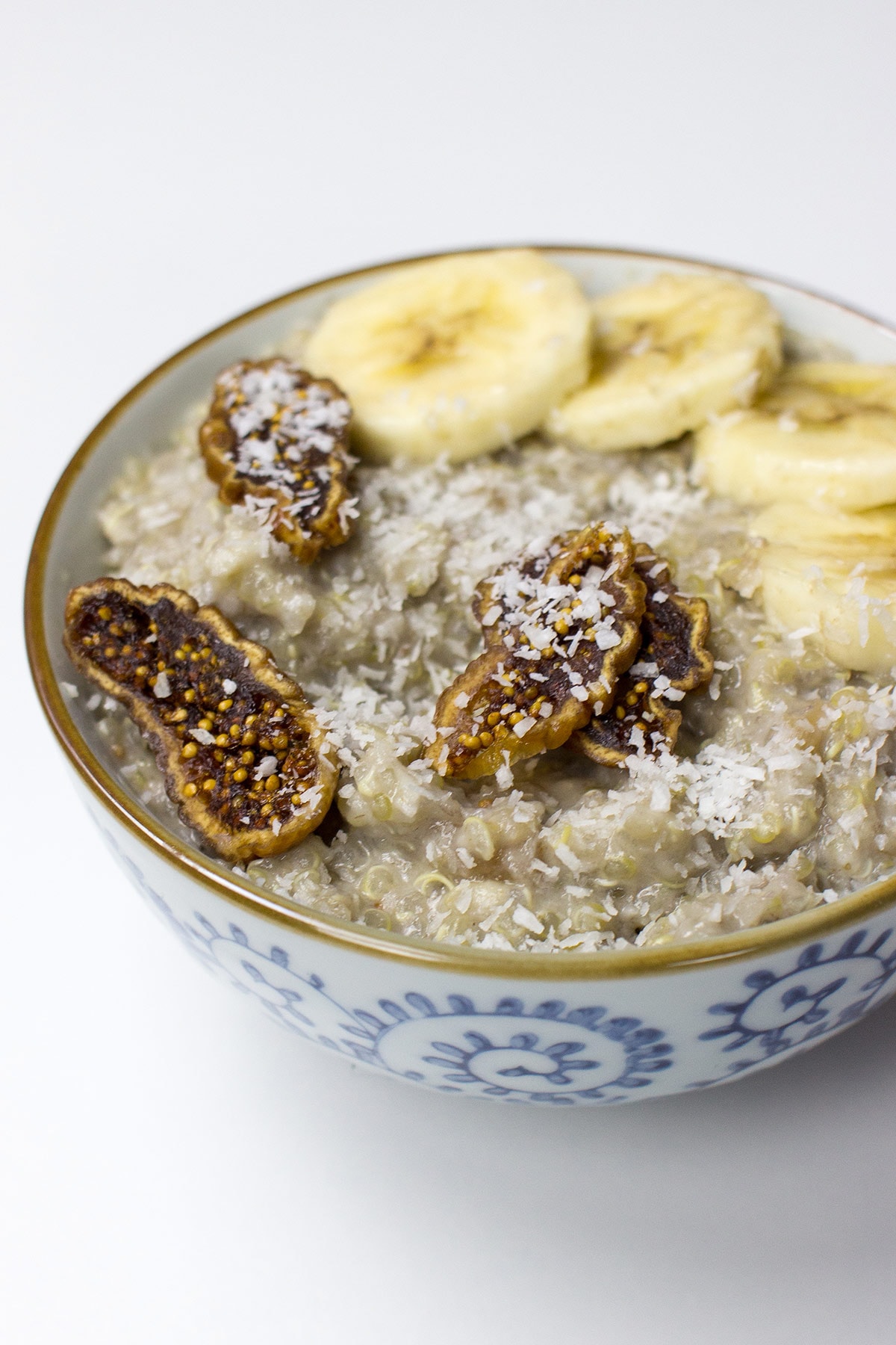A bowl of tropical quinoa breakfast porridge topped with banana slices, dried fig pieces, and shredded coconut on a white background.