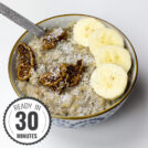 A bowl of tropical quinoa breakfast porridge topped with banana slices, dried fig pieces, and shredded coconut, served with a silver spoon on a white background with a timestamp in the left corner.