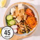 A birds-eye view of a marbled table with a wooden chopping board on top. On the board is a bowl of vegan bibimbap with a time stamp in the bottom left hand corner | Hurry The Food Up