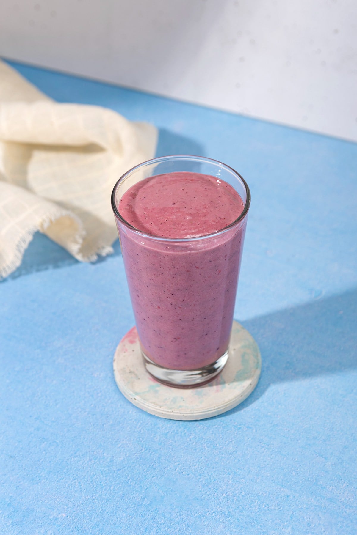 In the centre of a blue table is a white ceramic coaster with a tall glass full of blackberry smoothie. Next to the glass is a linen cloth | Hurry The Food Up