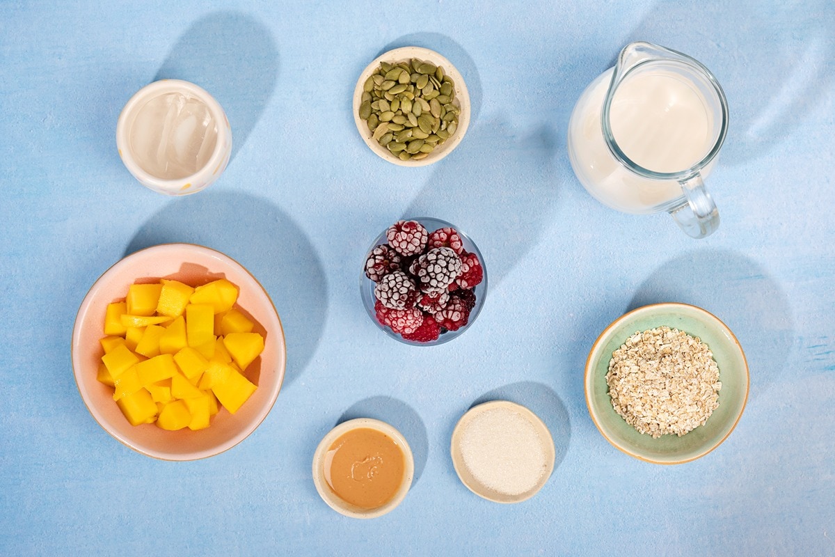 A birds-eye view of a blue table. On the table sits several bowls with the ingredients for a vegan blackberry smoothie. A bowl of mango cubes, ice cubes, almond butter, sugar, frozen blackberries, pumpkin seeds, oats, and a glass jug of soy milk | Hurry The Food Up