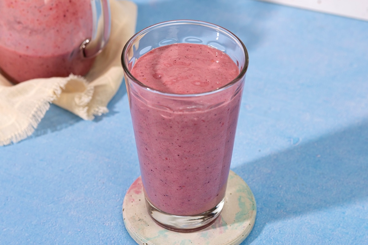 There is a blue table sat in front of a white stone wall. On the table is a white ceramic coaster with a tall glass full of blackberry smoothie. Behind the glass is a linen cloth with a glass jug containing more smoothie | Hurry The Food Up