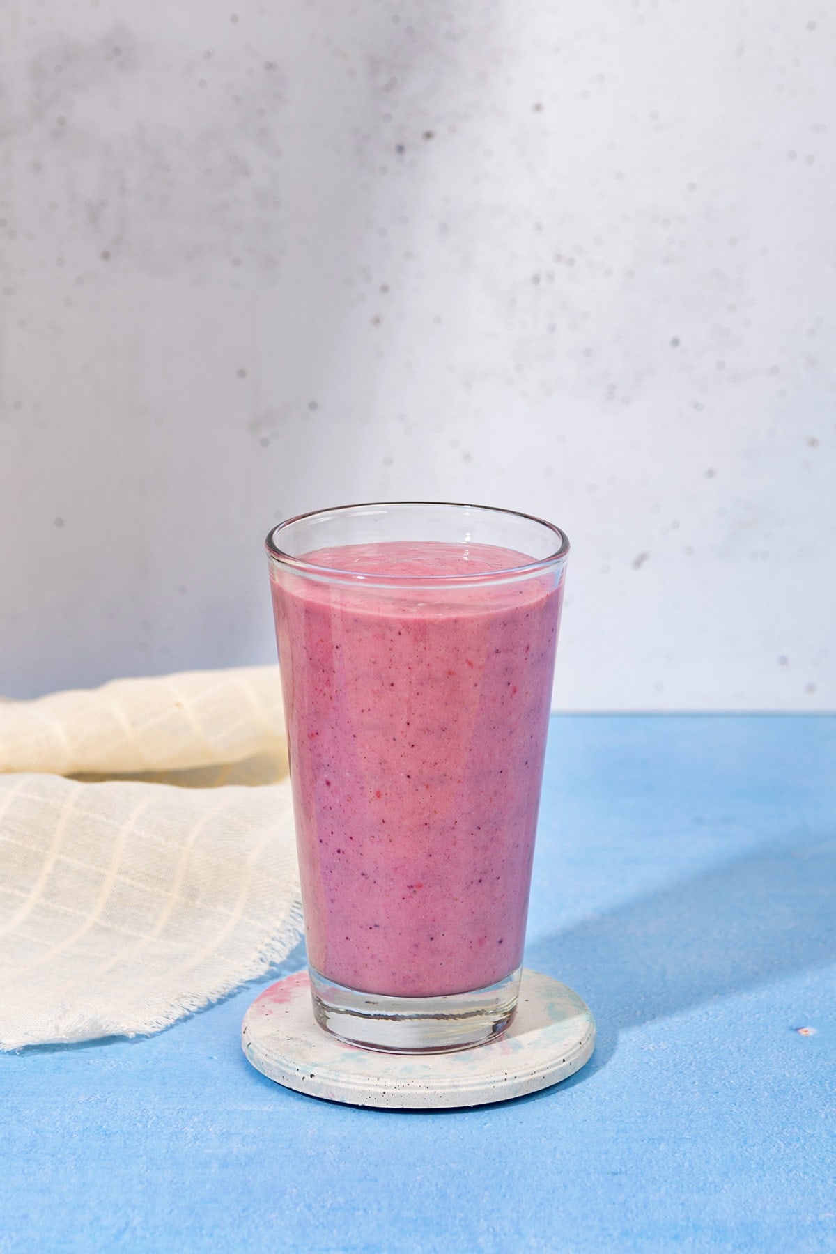 There is a blue table sat in front of a white stone wall. In the centre of the table is a white ceramic coaster with a tall glass full of blackberry smoothie. Next to the glass is a linen cloth | Hurry The Food Up