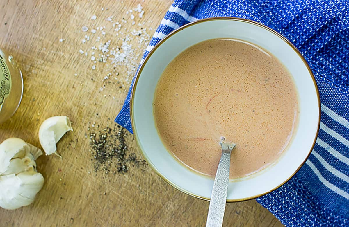 Caesar salad dressing in a bowl, few garlic gloves, some salt and pepper are on the table | Hurry The food Up