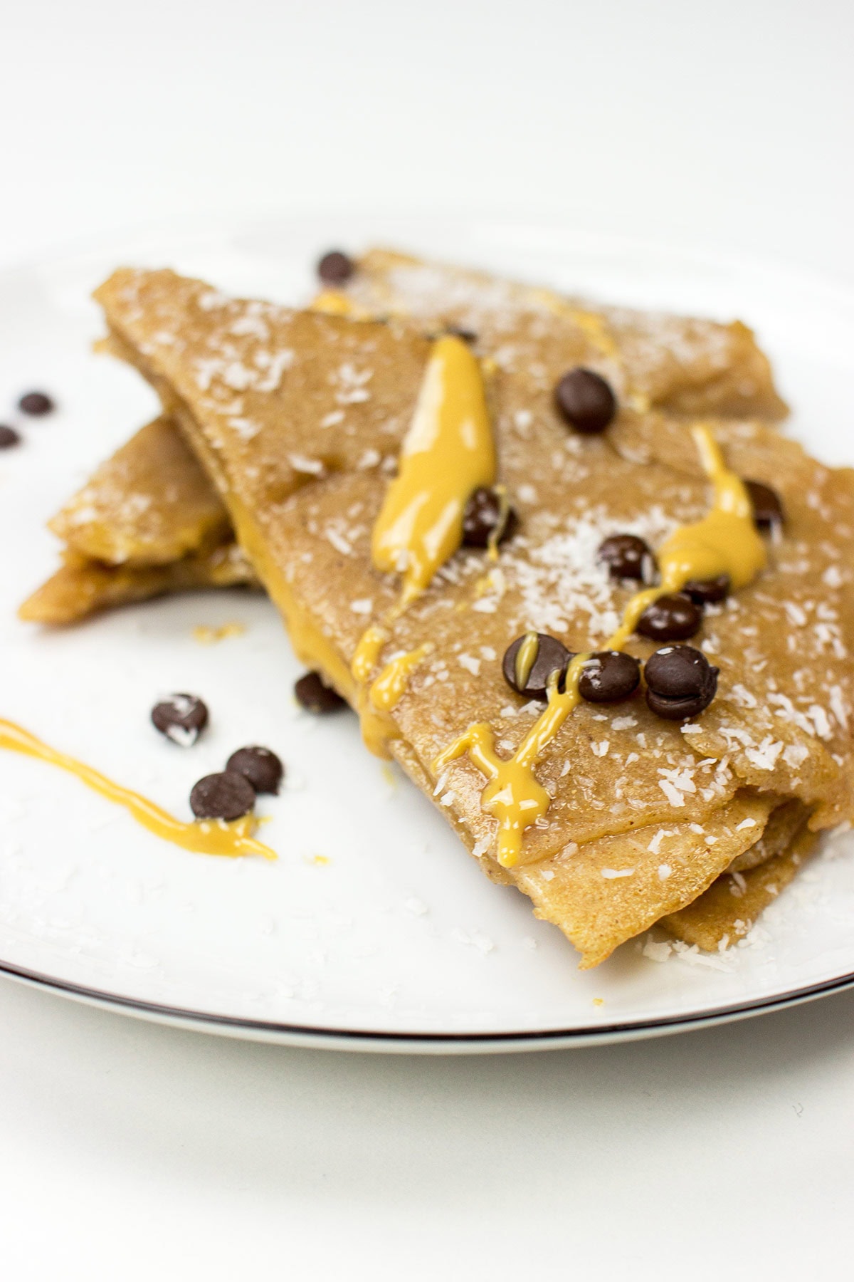 A white plate with two Vegan Chocolate & Coconut Crepes, topped with shredded coconut, chocolate chips, and drizzled with yellow sauce.