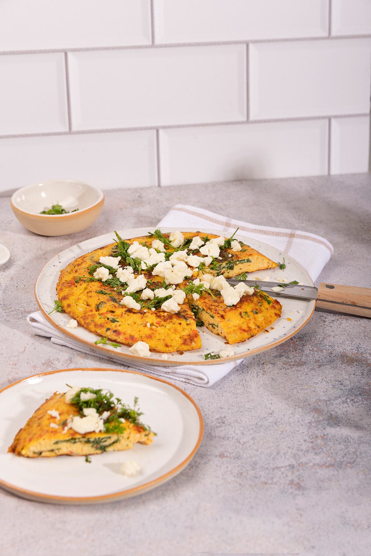 A plate of frittata sits on a white towel. There is a plate with a piece of frittata and a small bowl of herbs on the sides of it | Hurry The Food Up