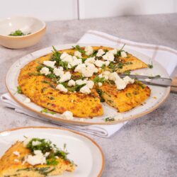 A plate of frittata sits on a white towel. There is a plate with a piece of frittata and a small bowl of herbs on the sides of it | Hurry The Food Up