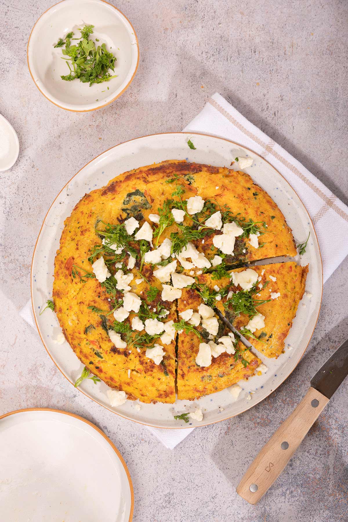 A birds-eye of Vegetable Frittata on a plate that is on a white towel. There is an empty plate and a small bowl of herbs on the sides of it | Hurry The Food Up