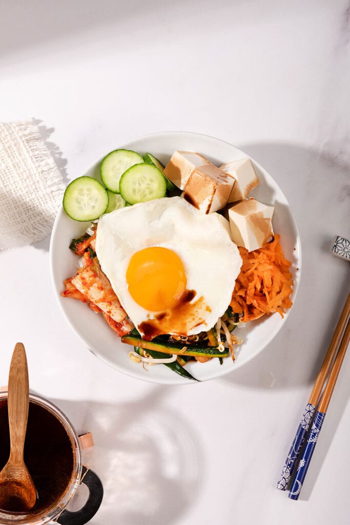 A birds-eye view of a vegetarian bibimbap in a large bowl. Underneath is a pot of soy sauce and a wooden spoon, to the right is a pair of chop sticks | Hurry The Food Up